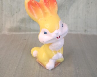 May include: A small yellow and white rabbit figurine with orange-tipped ears. The rabbit has blue eyes, a white muzzle, and a small red mouth. It is depicted in a seated position with its ears pointed upward, featuring a smooth, glossy finish.