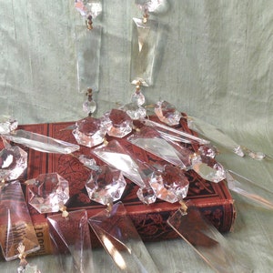 Op de afbeelding: Een verzameling van heldere glazen kristallen prisma's in verschillende vormen en maten, waaronder traanvormige, achthoekige en rechthoekige prisma's, gerangschikt op een rood boek met gouden letters.