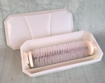 May include: A white plastic lint roller case with a hinged lid, containing a cylindrical roller with dense, light-colored bristles. The rectangular container features beveled edges and a clean, minimalist design for storing the cleaning tool.