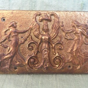 May include: A gold-toned metal plaque with three figures in relief. The central figure is winged and holds a staff. The figures are surrounded by ornate scrollwork.