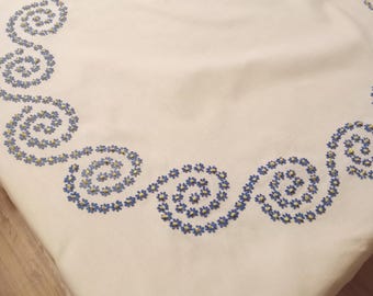 May include: Cream-colored tablecloth with a decorative border of blue and yellow floral embroidery. The embroidered design features a repeating pattern of swirling floral motifs around the edge of the cloth. The tablecloth is made of a soft, woven fabric.