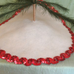 Può includere: Gonna per albero di Natale in feltro bianco con un bordo di paillettes rosse. Il bordo è fatto di piccole paillettes rotonde cucite insieme in un motivo continuo.