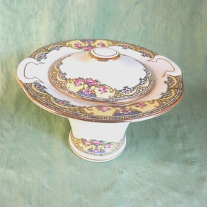 May include: A vintage, pedestal-style serving dish with a lid. The dish features a cream-colored base and lid, adorned with floral and gold trim. The design includes pink, yellow, and blue floral patterns, creating an elegant aesthetic.