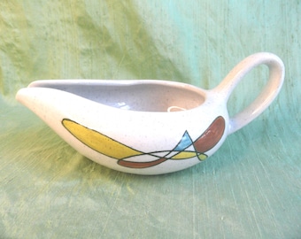 May include: White ceramic gravy boat with a speckled finish and a curved handle. The boat is decorated with an abstract design in yellow, brown, and blue. The gravy boat is designed for serving sauces and gravies.