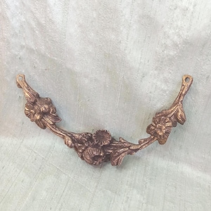 May include: A bronze-coloured decorative floral embellishment. The curved piece features detailed flowers and leaves, with small loops at each end. Likely for furniture or craft projects.