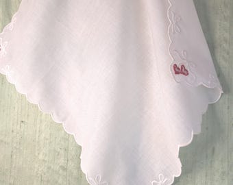 May include: A white square handkerchief with scalloped edges, featuring white embroidered bows and small red heart accents at the corners. A small rectangular tag is attached near the top center of the fabric.