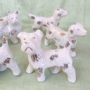 May include: A collection of white ceramic dog figurines with gold and brown accents. The dogs are in various poses, some standing and others sitting. The figurines have a glossy finish and are arranged on a light green surface.