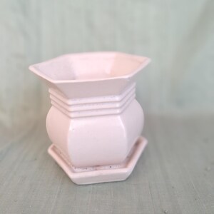 May include: A pale pink ceramic planter with a hexagonal shape and a matching hexagonal saucer base. The planter features a wide, flared rim and three horizontal ridges encircling the upper section, creating a distinct tiered profile.