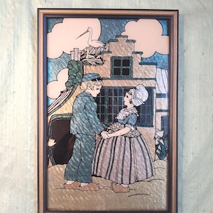 May include: Framed artwork depicting a couple in traditional Dutch attire, holding hands in front of a building. The artwork features a brown frame, with a blue and gold background. A stork is perched on the roof.