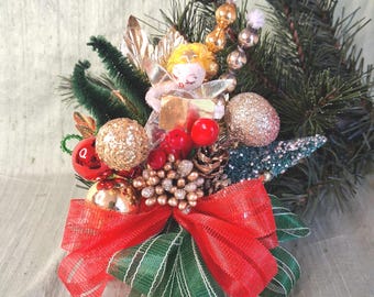 May include: A decorative Christmas arrangement featuring a small angel figurine with yellow hair, holding a gold book. The display includes red and gold glittered ornaments, pinecones, green pine branches, and a large red and green striped ribbon bow.