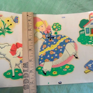 May include: Vintage children's sticker sheet featuring a girl in a blue dress, a white lamb, a red schoolhouse, and an ABC book. The sticker sheet is on a white background with a ruler for scale.