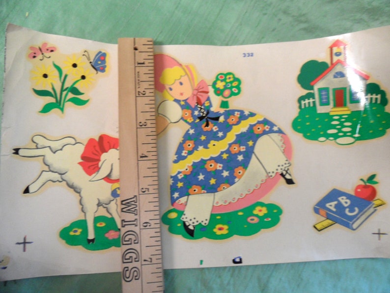 May include: Vintage children's sticker sheet featuring a girl in a blue dress, a white lamb, a red schoolhouse, and an ABC book. The sticker sheet is on a white background with a ruler for scale.