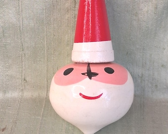 May include: A painted wooden ornament shaped like a round head with a pointed top. The figure features a white base, a pink face area, black circular eyes, a black nose, and a red curved mouth. A red and white conical hat sits on the head.