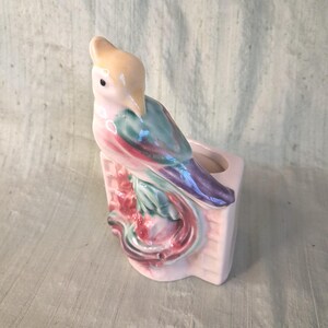 May include: A ceramic planter featuring a sculpted bird with a pale peach head and a body with blended teal, purple, and pink glazes. The bird is perched on a rectangular base with a grid pattern, and the piece has a glossy finish throughout.
