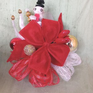 May include: A Christmas ornament with a red velvet bow, a small snowman figure, and various decorative elements. The ornament includes red and gold baubles, white mesh, and a glittery sphere, creating a cheerful holiday display.