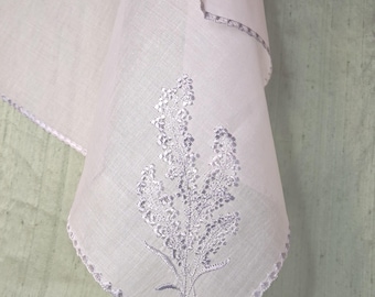 May include: A pale pink handkerchief featuring a delicate white floral embroidery pattern on one corner. The edges of the fabric are finished with a decorative light purple scalloped crochet border that frames the square textile piece.