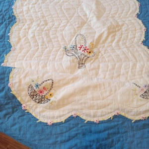 May include: A quilted, off-white table runner with a scalloped edge, embroidered with floral basket designs. The runner is set on a blue quilted surface. The edge is trimmed with small pink flowers.