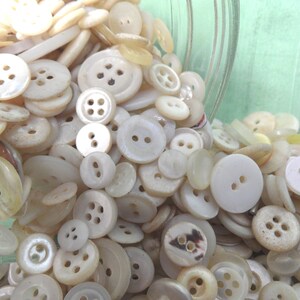 May include: A large collection of white and cream colored buttons of various sizes and styles. The buttons are spilling out of a glass jar.