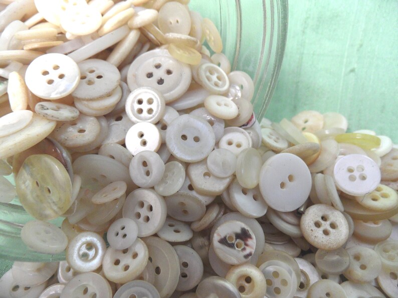 May include: A large collection of white and cream colored buttons of various sizes and styles. The buttons are spilling out of a glass jar.