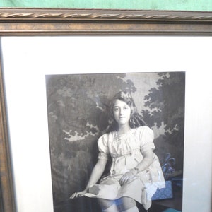 May include: A black and white portrait of a young girl wearing a white dress with a lace collar. She is sitting in a chair with a floral patterned background.