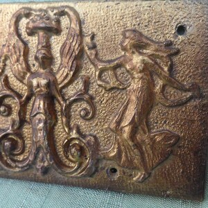 May include: A gold-toned metal plaque with a raised design of three figures. The figures are stylized and appear to be dancing. The plaque has a textured surface and is rectangular in shape.