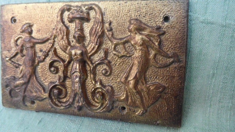 May include: A gold-toned metal plaque with a raised design of three figures. The figures are stylized and appear to be dancing. The plaque has a textured surface and is rectangular in shape.