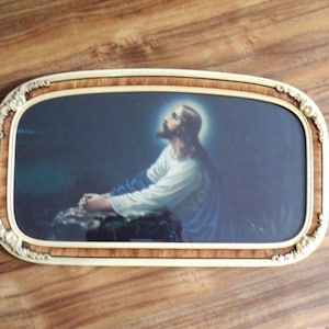 May include: A rectangular wall art piece featuring a depiction of a figure with long hair and a beard, kneeling in prayer with a glowing halo. The image is enclosed in a decorative frame with a wood-grain finish and ornate gold-toned corners.