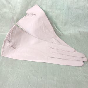 May include: A pair of pale pink leather gloves featuring a smooth finish, stitched finger detailing, and a decorative knotted loop accent at the wrist opening. The gloves are designed with a flared cuff and a clean, simple construction throughout.