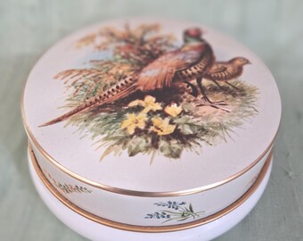 May include: A round white porcelain trinket box with a gold-trimmed lid featuring a colorful illustration of a pheasant in a grassy field with yellow flowers. The base of the box is decorated with small blue floral sprigs and gold accents.