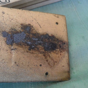 May include: A close-up of a rectangular metal plate with a gold finish. The plate has a dark brown stain running across the surface and several small holes.