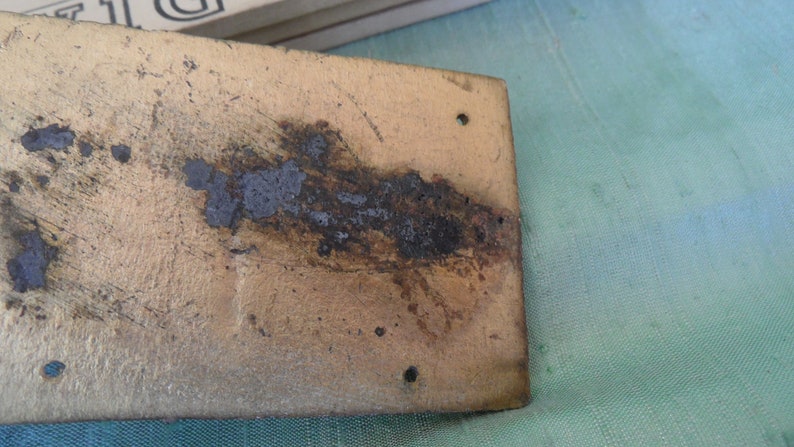 May include: A close-up of a rectangular metal plate with a gold finish. The plate has a dark brown stain running across the surface and several small holes.