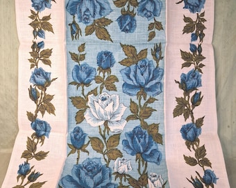May include: A rectangular textile featuring a central light blue panel decorated with blue and white rose blossoms and brown foliage. The panel is flanked by two vertical borders on a pale pink background, each displaying a continuous vine of blue roses.