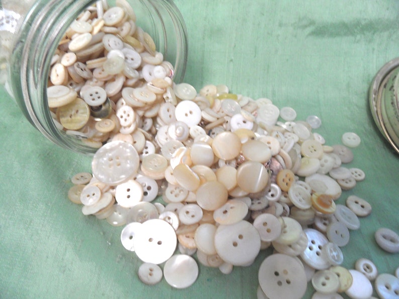 May include: A large collection of white and cream colored buttons of various sizes and shapes, spilling out of a glass jar onto a green surface.