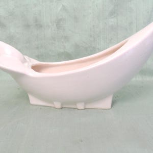 May include: A pale pink ceramic planter shaped like a stylised boat or vessel. The object features a smooth, matte finish with a pointed, elevated bow and a smaller, flared stern section. It rests on a rectangular base with small, integrated feet.