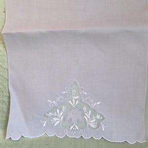 May include: A light lavender rectangular fabric runner featuring a scalloped bottom edge and a central white embroidered floral motif with cutwork details. The embroidery displays delicate leaves and blossoms centered on the lower section.
