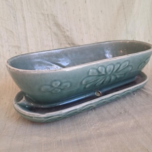 May include: An oval-shaped sage green ceramic planter with a matching drainage tray. The exterior of the planter features a raised floral relief pattern. The glazed finish has a glossy appearance, and the tray includes a small circular drainage hole.