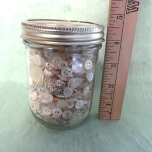 May include: A glass jar filled with a variety of white and cream colored buttons. The jar is approximately 4 inches tall and has a silver lid.