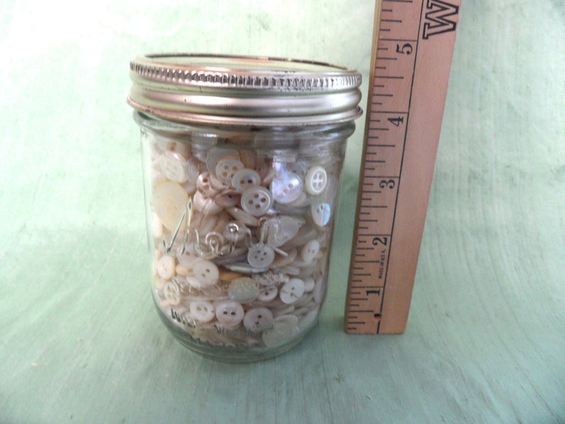 May include: A glass jar filled with a variety of white and cream colored buttons. The jar is approximately 4 inches tall and has a silver lid.