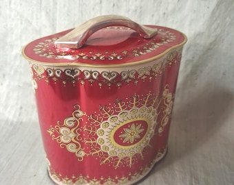 May include: An oval-shaped red metal storage tin with a matching lid and a silver-toned handle. The container features intricate gold and cream-colored decorative patterns, including a central circular medallion and ornate border designs.