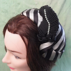 Könnte beinhalten: Ein schwarz-weiß gestreifter Turbanhut mit einer schwarzen Stoffblume und Perlenverzierung.