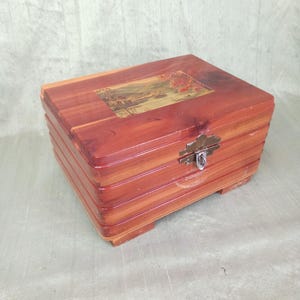 Può includere: Una scatola rettangolare in legno con coperchio incernierato. La scatola è realizzata in legno marrone rossiccio con un'immagine di paesaggio sul coperchio. Ha una chiusura con fermaglio in metallo e poggia su piedini. La scatola sembra essere vintage.