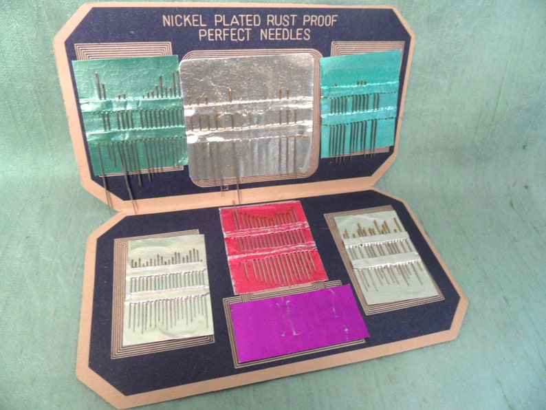 May include: A vintage cardboard box containing a collection of nickel-plated rust-proof sewing needles. The needles are organized in rows and are held in place by a green, silver, and red backing. The box has a black background and the text "Nickel Plated Rust Proof Perfect Needles" is printed on the front.
