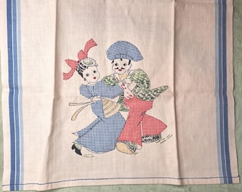 May include: A light pink rectangular kitchen towel featuring blue and white striped borders on the sides. The center displays an embroidered illustration of two figures dancing, wearing blue and red checkered clothing with a red bow accent.