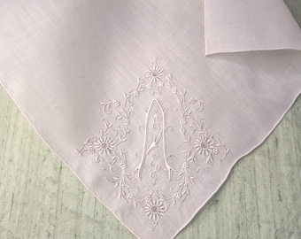 May include: A pale pink square handkerchief featuring white embroidered floral motifs surrounding a central letter A. The embroidery includes delicate vines, small blossoms, and leaves arranged in a diamond shape on the fabric surface.