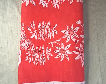 May include: A bright orange tea towel featuring a white botanical print of various flowers and seed pods. The bottom edge includes white text that reads, "flowers....designed and hand printed on linen." The fabric has a visible woven texture.