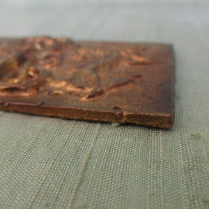May include: A small, rectangular, bronze-colored plaque with a raised, detailed design. The plaque has a slightly rough texture and appears to be made of metal.
