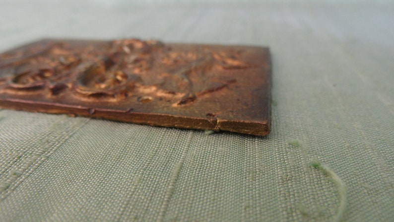 May include: A small, rectangular, bronze-colored plaque with a raised, detailed design. The plaque has a slightly rough texture and appears to be made of metal.