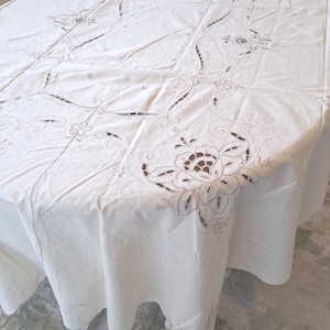 May include: A white tablecloth featuring intricate cutwork embroidery and detailed floral patterns. The fabric displays delicate needlework with openwork sections forming scrolling vines and large blossoms that drape over the edges of the table.