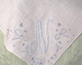 May include: A white linen handkerchief featuring a light blue embroidered monogram letter N in the center, surrounded by delicate floral motifs, small dots, and decorative stitching along the scalloped edge of the fabric corner.