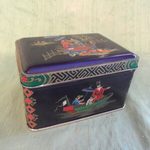 Può includere: Una scatola vintage in latta blu e oro con un design di ispirazione asiatica. Il coperchio presenta una scena di persone in abiti tradizionali su una barca. Il fondo della scatola presenta una scena simile con una barca e persone.